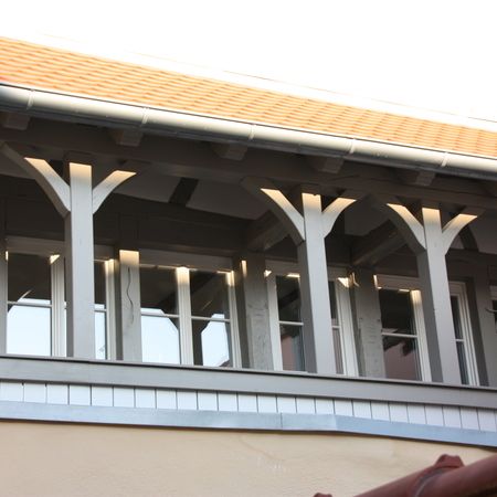Teilweise verglaste Veranda mit grauer Holzkonstruktion und Zierstreben unterhalb eines roten Ziegeldachs.