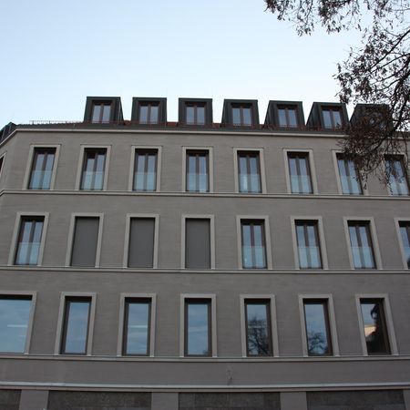 Mehrstöckiges Gebäude mit heller Fassade, rechteckigen Fenstern und Dachgauben, symmetrisch angeordnet.