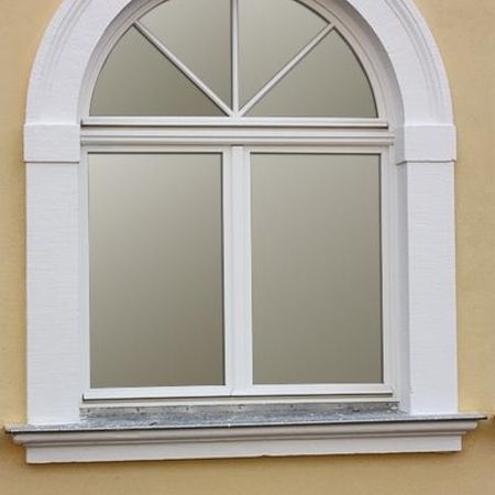 Rundbogenfenster mit weißem Rahmen und dekorativem Scheibenkreuz in gelber Putzfassade.