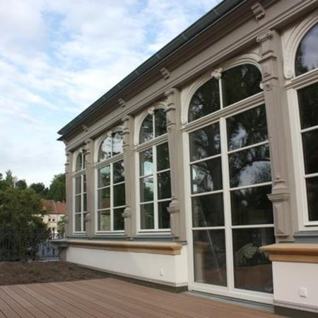 Großzügige Fensterfront in historischer Fassade mit rundbogigen Holzfenstern und klassizistischen Stuckelementen.