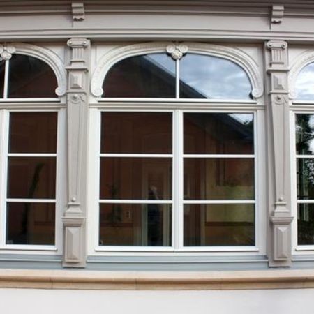 Nahaufnahme historischer weißer Holzfenster mit rundbogigem Abschluss und Zierverglasung, eingefasst in kunstvolle Stuckrahmen.