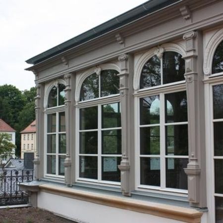 Außenansicht eines Wintergartens mit historischen, weißen Rundbogenfenstern und reich verzierten Fassadenelementen.