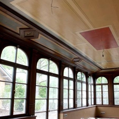Innenansicht eines Wintergartens mit hohen Rundbogenfenstern und Holzrahmen, Tageslicht fällt in den Raum.