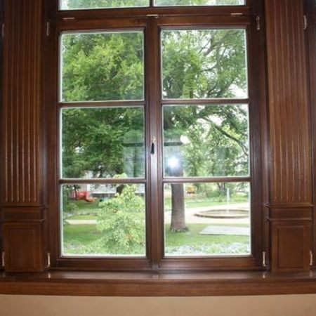 Innenansicht eines rechteckigen Holzfensters mit aufgesetzten Sprossen und profiliertem Holzrahmen, Ausblick auf Bäume und eine Rasenfläche mit Brunnen.