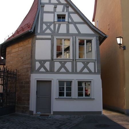 Schmales Fachwerkhaus mit steil zulaufendem Satteldach, grauen Holzfenstern und grauer Hauseingangstür, umgeben von Kopfsteinpflaster und seitlich angrenzenden Gebäuden.