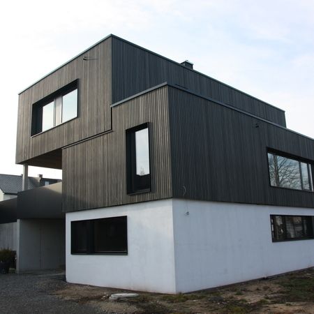 Modernes Einfamilienhaus mit Holzfassade: Zweigeschossiges Gebäude mit vertikaler Holzverkleidung und großen, schwarz gerahmten Fenstern.