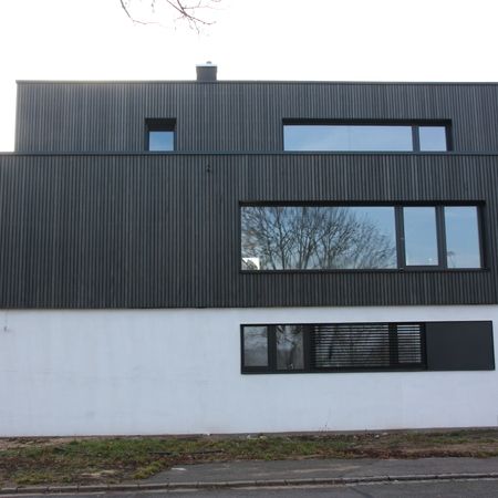 Moderne Fassade mit großem Fensterband: Flaches Gebäude mit dunkler Holzverkleidung im oberen Bereich und weißem Sockel, mittig ein breites Fensterband.