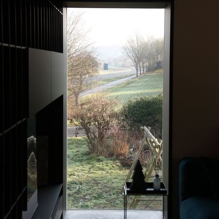 Schmales Panoramafenster im Innenraum: Blick durch ein raumhohes Fenster auf eine leicht ansteigende Straße mit Büschen im Vordergrund.