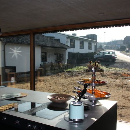 Küchensituation mit großem Fenster: Blick aus einer modernen Küche mit Kochinsel durch ein großes Fenster auf eine Einfahrt und ein gegenüberliegendes Haus.