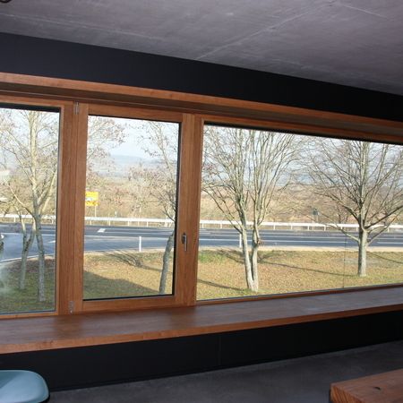 Großes Holzfenster mit Fensterbank: Innenansicht eines breiten Fensters mit Holzrahmen, Blick auf Straße mit Leitplanke und kahlen Bäumen.