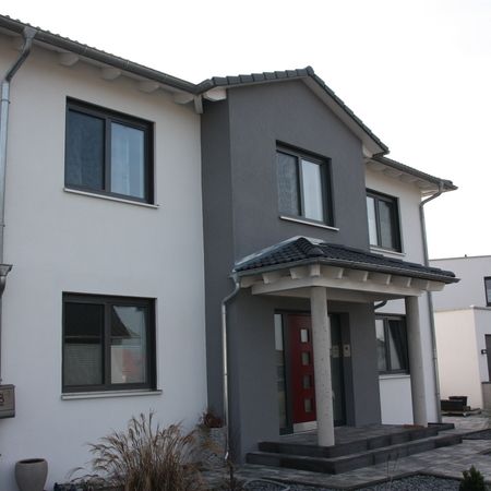 Zweifamilienhaus mit roter Eingangstür: Vorderansicht eines modernen Hauses mit Putzfassade in Grau-Weiß, zentraler Eingang und rechteckige Fenster mit dunklen Rahmen.