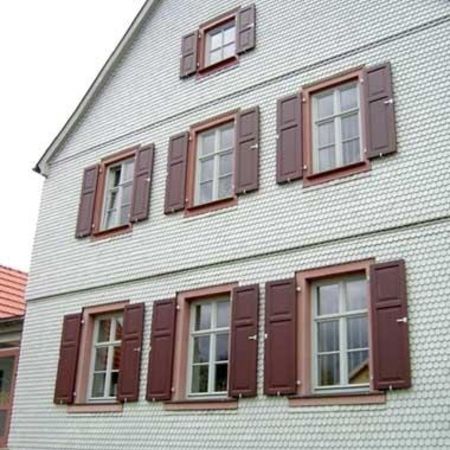 Ein dreistöckiges Haus mit einer Fassadenverkleidung aus kleinen, gleichmäßigen Schindeln in hellem Grau. Die Fenster sind symmetrisch angeordnet und besitzen klassische weiße Sprossenrahmen.