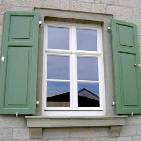 Ein einzelnes Fenster mit weißem Sprossenrahmen, eingefasst in eine hellgraue Steinfassade. Das Fenster wird von zwei grünen Fensterläden flankiert, die geöffnet sind.