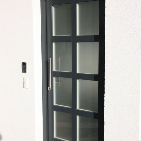 Dunkelgraue Haustür mit Glasrahmen: Schmale Haustür mit satiniertem Glas in rechteckigem Rastersystem, klarer Griffleiste und moderner Gegensprechanlage.