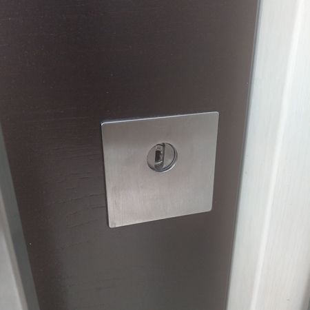 Nahaufnahme eines eckigen, flächenbündig eingelassenen Schlüssellochs aus Edelstahl in einer dunklen Haustür.