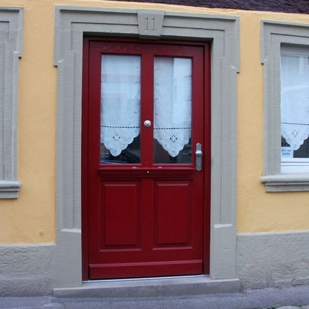 Rote Haustür mit Klarglas und Vorhang: Zweiflügelige, rote Holzhaustür mit Verglasung und weißem Spitzenvorhang, eingefasst in hellgrauen Sandsteinrahmen.