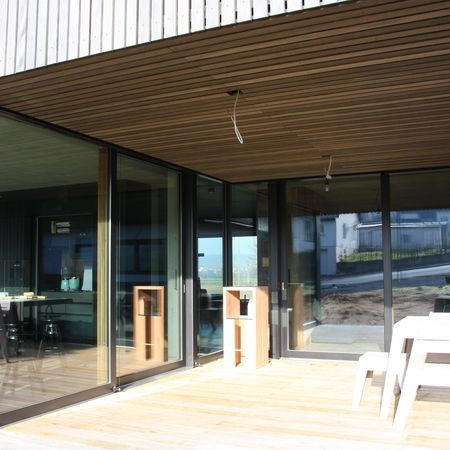 Großzügige, bodentiefe Hebeschiebetür mit dunklem Aluminiumrahmen in einer modernen Holzfassade, öffnet sich zu einer Holzterrasse mit Sitzgruppe.