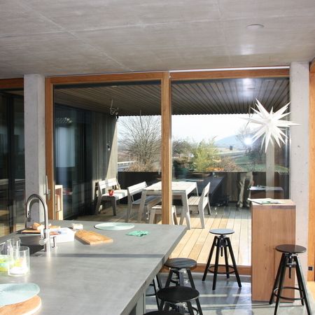 Blick aus einer modernen Küche auf eine überdachte Terrasse mit Esstisch, durch eine Hebeschiebetür mit Holzrahmen und klarer Verglasung.