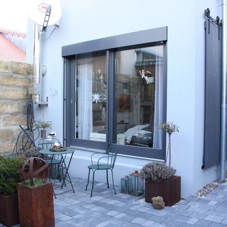 Graue Hebeschiebetür mit integrierter Fensterfunktion öffnet sich zu einer kleinen Terrasse mit Tisch und zwei Stühlen, flankiert von Pflanzenkübeln.