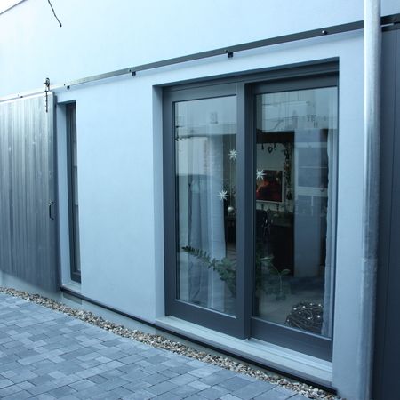 Zweiflügelige Hebeschiebetür in anthrazitfarbenem Rahmen, eingebaut in eine weiße Putzfassade, mit oben montierter Laufschiene für Schiebeläden.