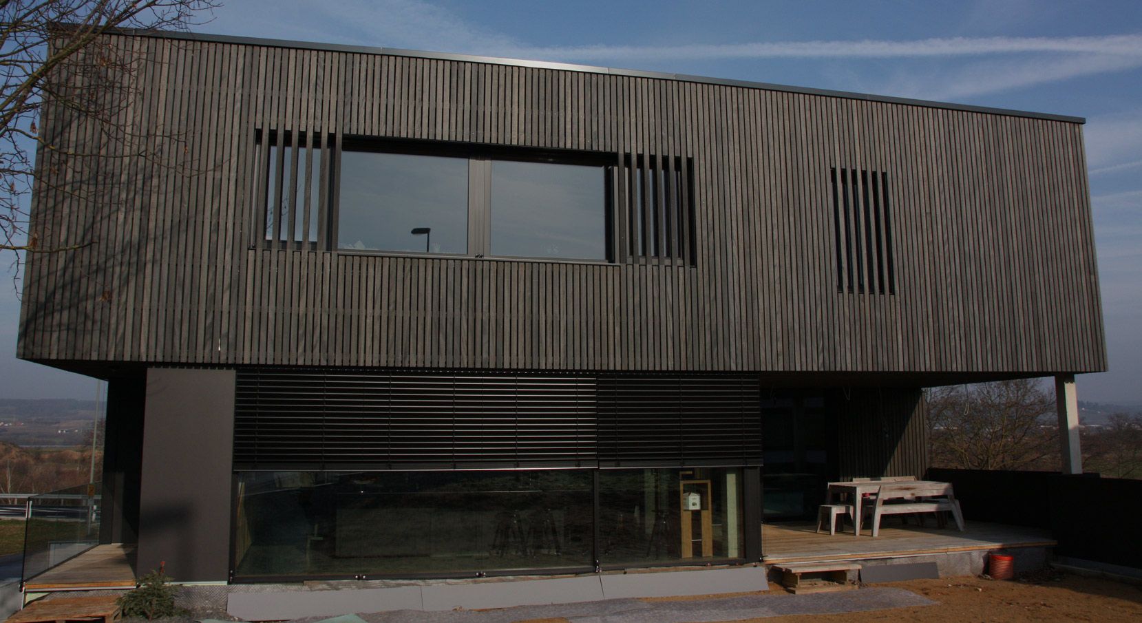 Modernes Wohnhaus mit dunkler, vertikaler Holzfassade und großen Fensterflächen, teils mit Lamellen oder Glasfront im Erdgeschoss.